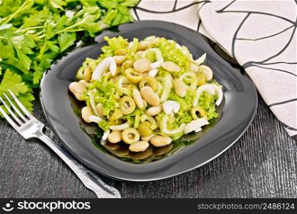 Salad of beans, olives, stalk celery, onion and lettuce, dressed with vegetable oil in a plate, napkin and fork on black wooden board background