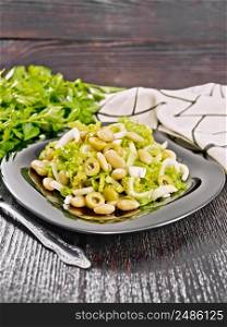 Salad of beans, olives, stalk celery, onion and lettuce, dressed with vegetable oil in a plate, towel and fork on dark wooden board background