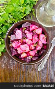 Salad of beets and potatoes, seasoned with vegetable oil and vinegar in a bowl, parsley and a fork on the background of a dark wooden board on top