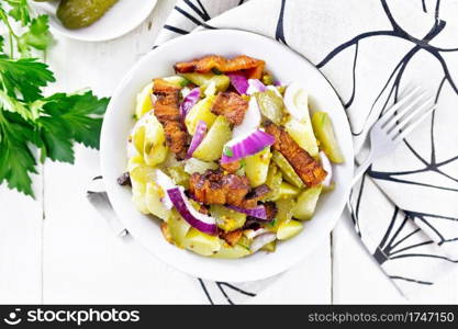 Salad of boiled potatoes, fried bacon, red onions and pickled cucumbers, seasoned with grain mustard, spices and vegetable oil in a plate on napkin against wooden board from above