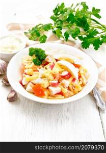 Salad of crab sticks, cheese, garlic, tomatoes and eggs with mayonnaise in a plate, kitchen towel, parsley and fork on a light wooden board background