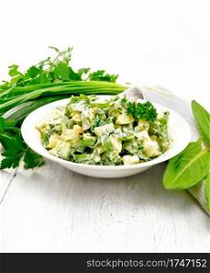 Salad of cucumber, sorrel, boiled potatoes, eggs and herbs, dressed with mayonnaise in a white plate, parsley, green onions and kitchen towel against the background of light wooden board