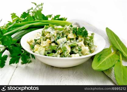 Salad of cucumber, sorrel, boiled potatoes, eggs and herbs, dressed with mayonnaise in a white plate, parsley, green onions and towel against the background of wooden boards