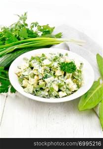 Salad of cucumber, sorrel, boiled potatoes, eggs and herbs, dressed with mayonnaise in a white plate, parsley, green onions and kitchen towel on wooden board background