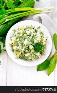 Salad of cucumber, sorrel, boiled potatoes, eggs and herbs, dressed with mayonnaise in a white plate, parsley, green onions and kitchen towel on background of wooden board on top