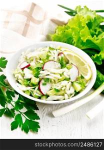 Salad of fresh cabbage, radish, lettuce, cucumber and green onion, dressed with vegetable oil in plate, napkin and parsley on the background of light wooden board