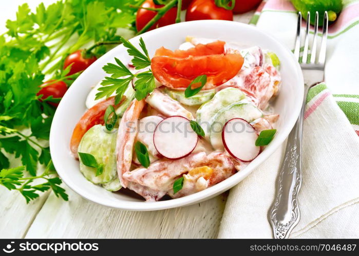 Salad of fresh tomatoes, cucumbers and radish with green onions and parsley, flavored with mayonnaise and sour cream in a plate, napkin and fork on a wooden plank background