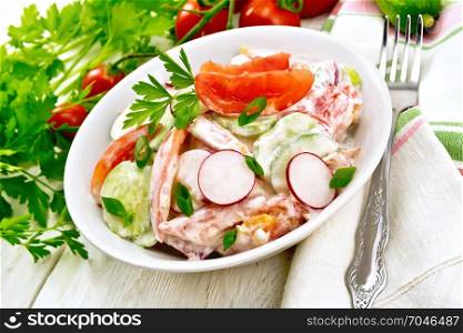 Salad of fresh tomatoes, cucumbers and radish with green onions and parsley, flavored with mayonnaise and sour cream in a plate, napkin and fork on a wooden plank background