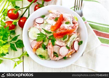 Salad of fresh tomatoes, cucumbers and radish with green onions and parsley, flavored with mayonnaise and sour cream in a plate, towel and fork on a wooden board background on top