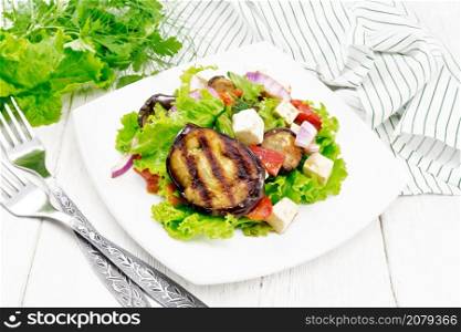 Salad of fried eggplant, fresh tomato with red onion and salted feta cheese, seasoned with vegetable oil and soy sauce on lettuce in plate, towel, fork and parsley on wooden board background