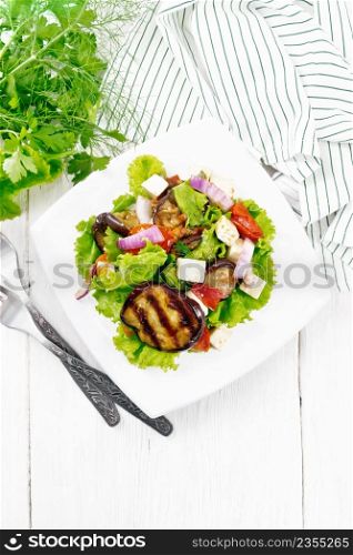Salad of fried eggplant, fresh tomato with red onion and salted feta cheese, seasoned with vegetable oil and soy sauce on lettuce in plate, towel, parsley on wooden board background from above