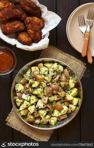 Salad of jacket potato, red onion and herbs with homemade breaded and fried chicken wings, photographed overhead on dark wood with natural light