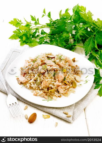 Salad of smoked salmon, rice, sunflower and pumpkin seeds, almonds, seasoned with honey and olive oil in a plate on towel, parsley and fork on background of light wooden board