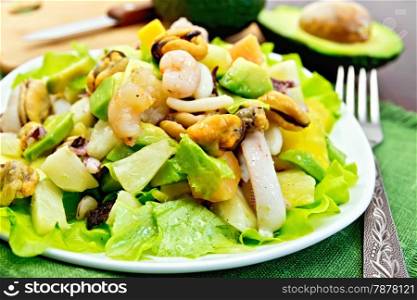 Salad with shrimps, octopus, mussels and calamari with avocado, lettuce, pineapple in a plate on a napkin, fork, knife on the background dark wooden boards
