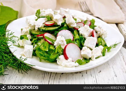 Salad with spinach, cucumbers, radish and salted cheese, dill and green onions in a bowl, towel on the background of a light wooden board