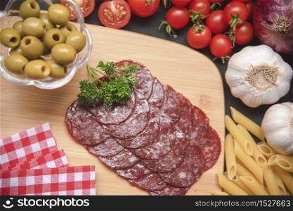 Salami Italian ingredient food on wooden cutting board on black table. Red Tomato with Salami, garlic, onion, green olive and pasta