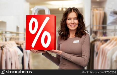sale and business concept - happy female shop assistant or saleswoman holding shopping bag with percentage sign over clothing store background. saleswoman with percentage sign on shopping bag