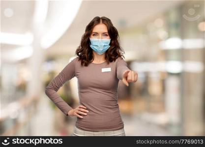 sale and pandemic concept - happy female shop assistant or saleswoman in medical mask with name tag pointing finger to camera over shopping mall background. female shop assistant in mask pointing to camera