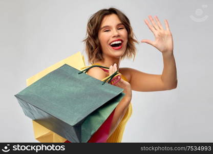 sale and people concept - happy smiling young woman in mustard yellow top and jeans with shopping bags waving hand over grey background. happy smiling young woman with shopping bags