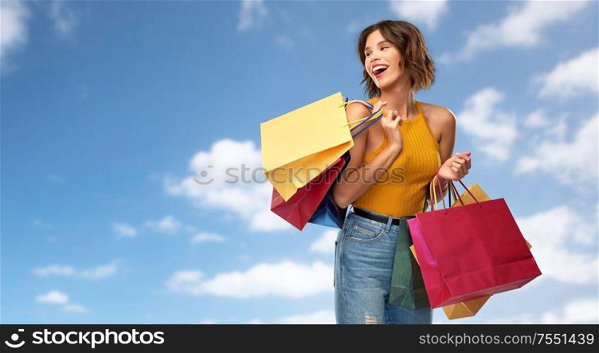 sale and people concept - happy smiling young woman in mustard yellow top and jeans with shopping bags over blue sky and clouds background. happy smiling young woman with shopping bags