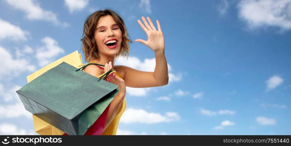 sale and people concept - happy smiling young woman in mustard yellow top and jeans with shopping bags waving hand over blue sky and clouds background. happy smiling young woman with shopping bags