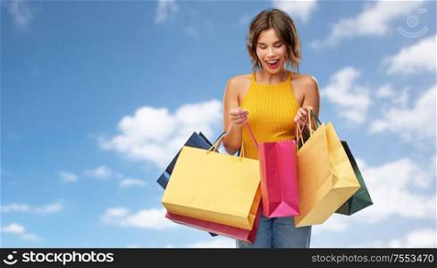 sale and people concept - happy smiling young woman in mustard yellow top and jeans with shopping bags over blue sky and clouds background. happy smiling young woman with shopping bags