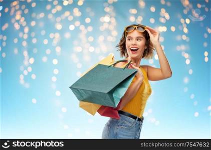 sale and people concept - happy smiling young woman in yellow top and jeans with shopping bags over festive lights on blue background. happy smiling young woman with shopping bags