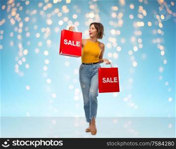 sale and people concept - happy smiling young woman in yellow top and jeans with shopping bags over holidays lights on blue background. happy smiling young woman with shopping bags
