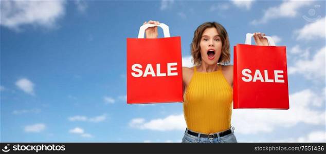 sale and people concept - surprised young woman in mustard yellow top and jeans with shopping bags , over blue sky and clouds background. surprised young woman with shopping bags on sale
