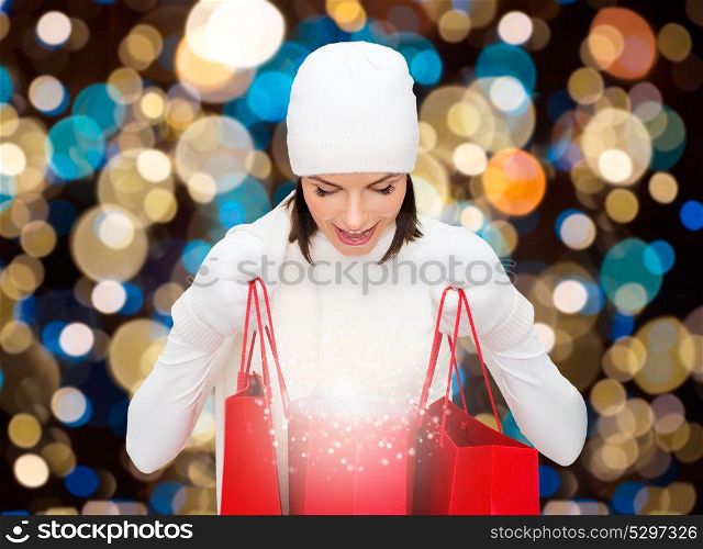 sale, christmas, holidays and people concept - happy woman in winter hat looking to fairy dust in shopping bags over lights background. woman in winter hat with christmas shopping bags