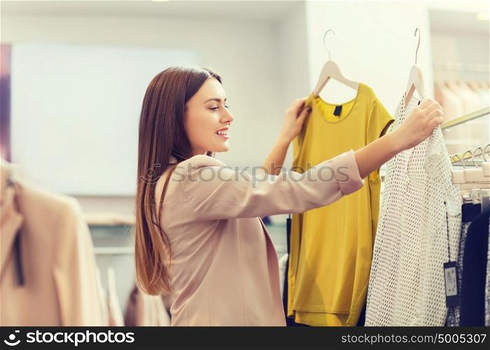 sale, clothes , shopping, fashion and people concept - happy young woman choosing between two shirts in mall or clothing store. happy young woman choosing clothes in mall
