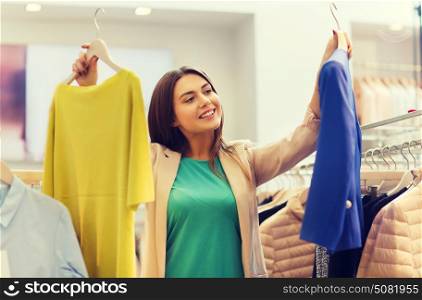 sale, clothes , shopping, fashion and people concept - happy young woman choosing between shirt and jacket at clothing store. happy young woman choosing clothes in mall