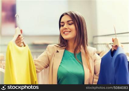 sale, clothes , shopping, fashion and people concept - happy young woman choosing between shirt and jacket at clothing store. happy young woman choosing clothes in mall. happy young woman choosing clothes in mall