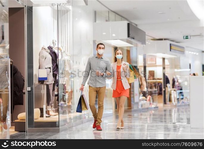 sale, consumerism and pandemic concept - happy young couple wearing face protective medical mask for protection from virus disease with shopping bags walking in mall. couple in medical masks with shopping bags in mall