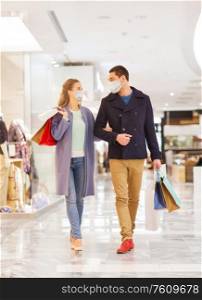 sale, consumerism and pandemic concept - happy young couple wearing face protective medical mask for protection from virus disease with shopping bags walking and talking in mall. couple in medical masks with shopping bags in mall