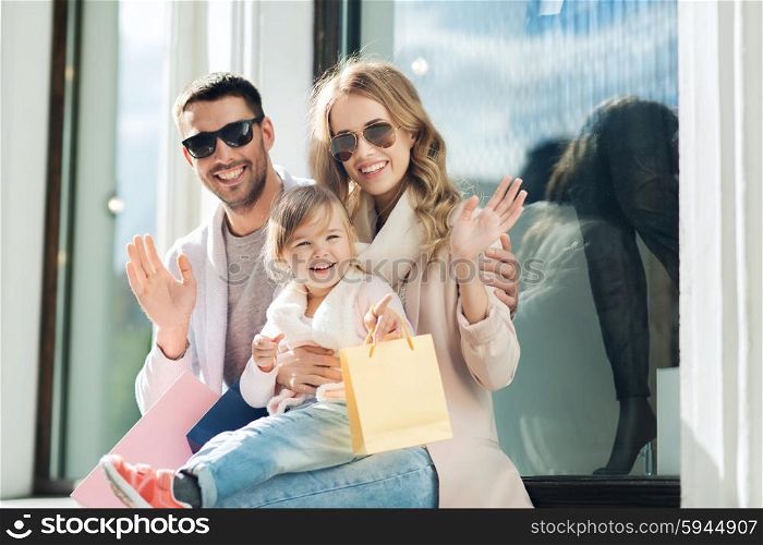 sale, consumerism and people concept - happy family with little child and shopping bags waving hands at shop window in city