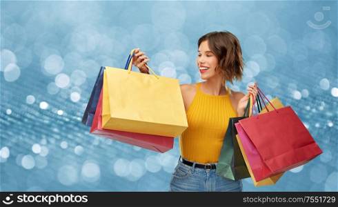 sale, consumerism and people concept - happy smiling young woman in mustard yellow top and jeans with shopping bags over shimmering blue background. happy smiling young woman with shopping bags