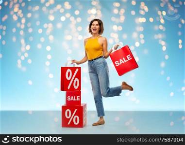 sale, consumerism and people concept - happy smiling young woman in mustard yellow top and jeans with shopping bags over holidays lights on blue background. happy smiling young woman with shopping bags