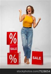 sale, consumerism and people concept - happy smiling young woman in sunglasses, mustard yellow top and jeans with shopping bags celebrating success over grey background. happy smiling young woman with shopping bags