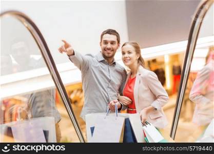 sale, consumerism and people concept - happy young couple with shopping bags rising on escalator and pointing finger in mall
