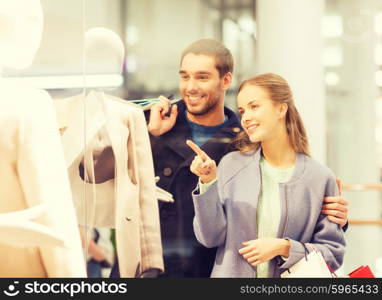 sale, consumerism and people concept - happy young couple with shopping bags pointing finger to shop window in mall