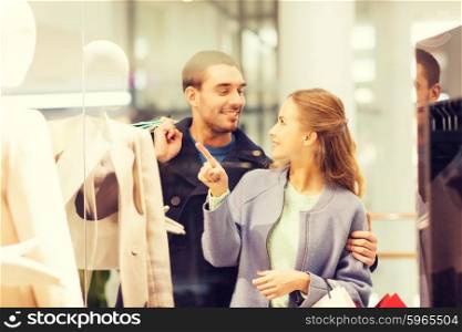 sale, consumerism and people concept - happy young couple with shopping bags pointing finger to shop window in mall