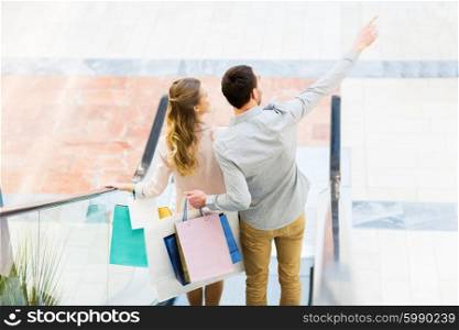 sale, consumerism and people concept - happy young couple with shopping bags going down by escalator and pointing finger in mall