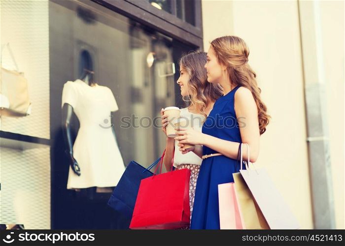 sale, consumerism and people concept - happy young women with shopping bags and coffee paper cups looking at shop window outdoors
