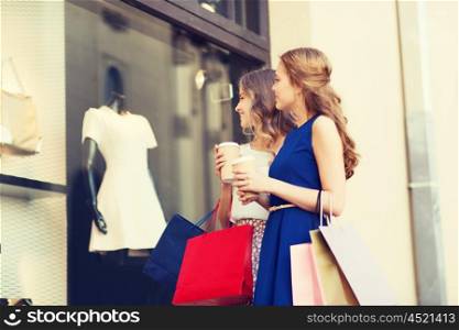 sale, consumerism and people concept - happy young women with shopping bags and coffee paper cups looking at shop window outdoors