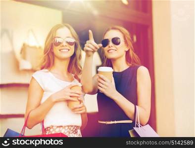 sale, consumerism and people concept - happy young women with shopping bags and coffee paper cup pointing finger at shop window in city. young women with shopping bags and coffee at shop