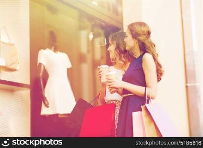 sale, consumerism and people concept - happy young women with shopping bags and coffee paper cups looking at shop window outdoors. young women with shopping bags and coffee at shop