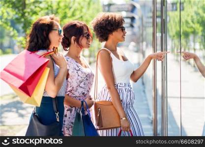 sale, consumerism and people concept - happy young women with shopping bags pointing finger to shop window in city. women with shopping bags looking at shop window