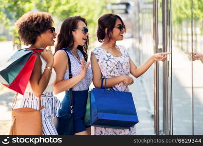 sale, consumerism and people concept - happy young women with shopping bags pointing finger to shop window in city. women with shopping bags looking at shop window