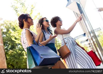 sale, consumerism and people concept - happy young women with shopping bags pointing finger to shop window in summer city. women with shopping bags looking at shop window